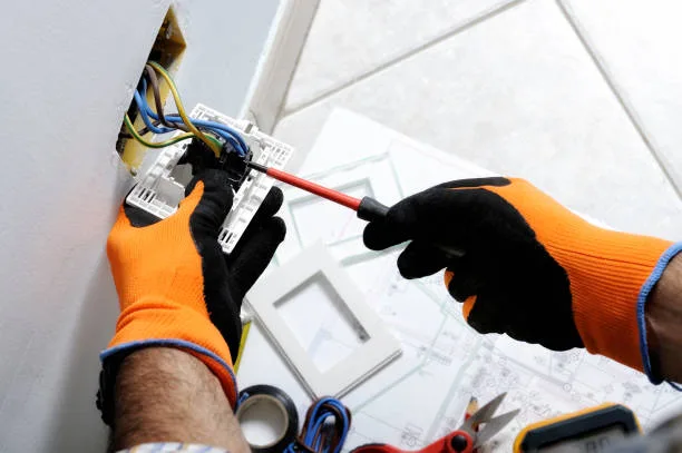 Gloved hands screwdriving a switch mechanism into a wall box beside a wiring plan and tools