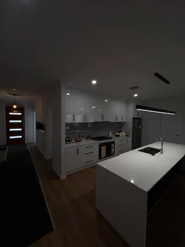 Downlights on over a stone island in a white kitchen with the hallway dark beyond