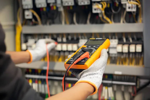 Gloved hands holding a multimeter across the terminals of an industrial distribution board
